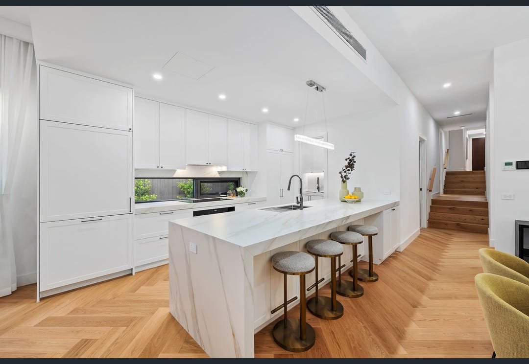 White kitchen with a veined island benchtop, breakfast stools and herringbone timber flooring.